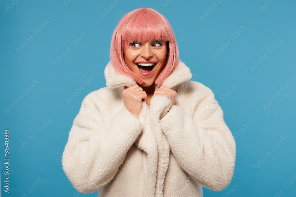 Overjoyed attractive young female with short pink bob haircut looking exitedly aside with wide eyes and mouth opened, holding colar of her white coat while posing over blue background Overjoyed attractive young female with short pink bob haircut looking exitedly aside with wide eyes and mouth opened, holding colar of her white coat while posing over blue background