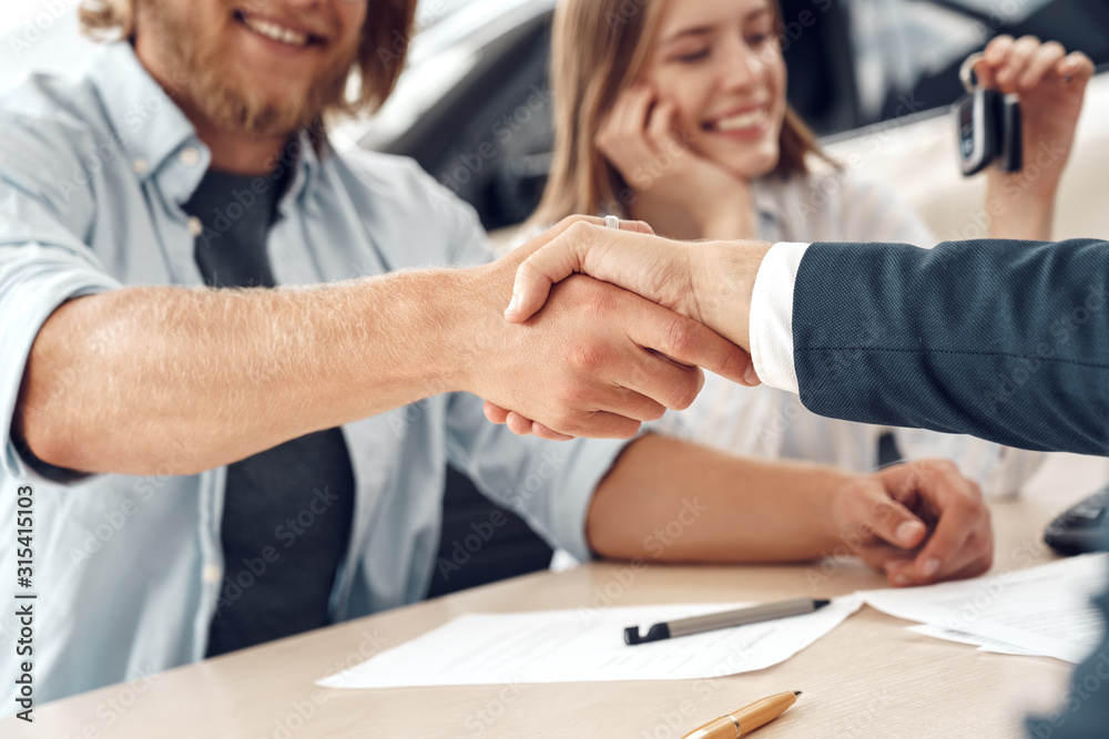 Obraz premium Car Salon. Young couple sitting at table man shaking hands with agent making agreement close-up blurred backgound smiling cheerful