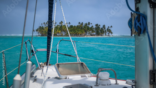 A beautiful Deserted Tropical Island overgrown with green Palm Trees seen from a white Sailboat surrounded by Turquoise Water in the San Blas Islands, Panama.