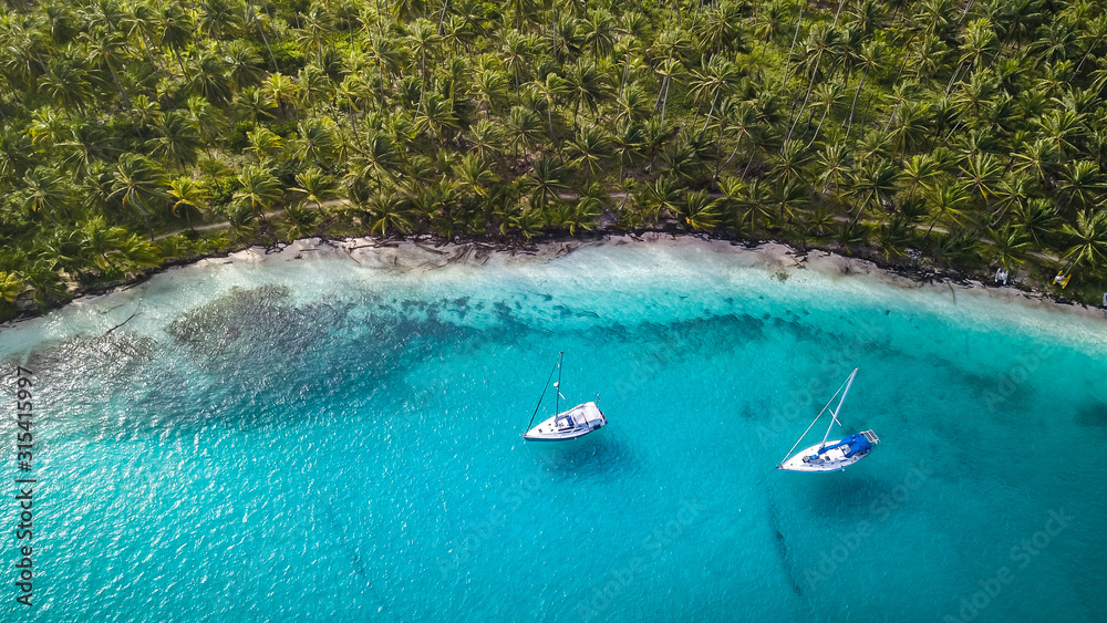 San Blas Islands, Panama - Aerial Drone Top Down View of two Sailing Yachts anchored in Turquoise Water right next to perfect White Sand Beach of Caribbean Tropical Island full of green Palm Trees. - obrazy, fototapety, plakaty 