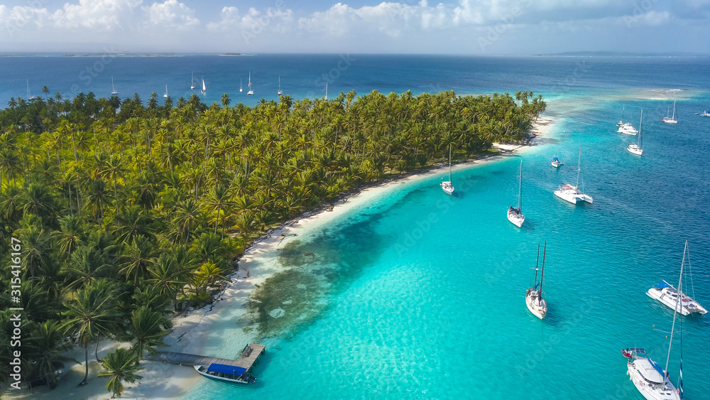 San Blas Islands, Panama - Drone Aerial View of many Sailboats & Sailing Yachts anchored in turquoise Water of Blue Lagoon next to white Sand Beach of Tropical Caribbean Island with green Palm Trees.