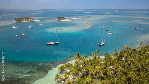 San Blas Islands, Panama - The Sailing Paradise! A beautiful Drone Shot of many Sailboats anchored in a Blue Lagoon between some Tropical Caribbean Islands overgrown with green Palm Trees.
