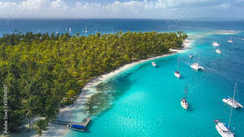 San Blas Islands, Panama - Drone Aerial View of many Sailboats & Sailing Yachts anchored in turquoise Water of Blue Lagoon next to white Sand Beach of Tropical Caribbean Island with green Palm Trees. photo