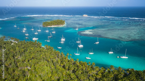 San Blas Islands, Panama - The Sailing Paradise! Aerial Drone Shot of many Sailboats anchored in Blue Lagoon next to Tropical Island with green Palm Trees surrounded by Coral Reefs in Caribbean Sea.