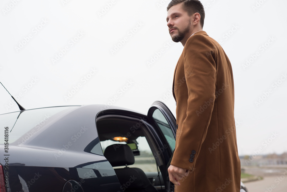 Portrait of a young guy in business clothes, a businessman of thirty years opens the car door to exit. In an interesting authoring