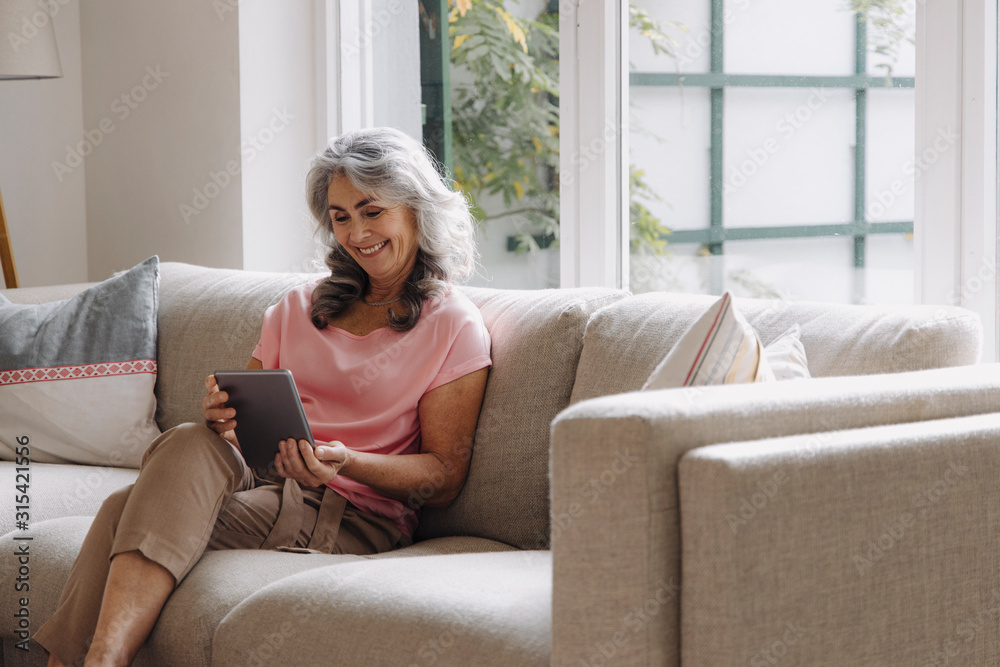 Smiling mature woman using tablet sitting on couch at home Smiling mature woman using tablet sitting on couch at home