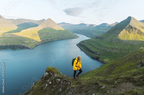 Lonely tourist in yellow jacket looking over majestic fjords of Funningur, Eysturoy island, Faroe Islands. Landscape photography