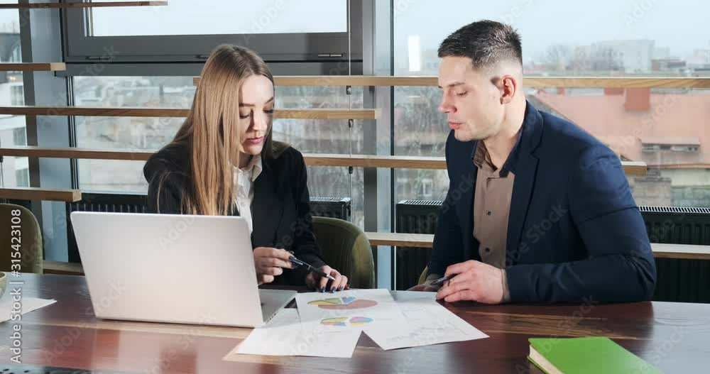 Man and a woman discussing work in the brightly lit modern office. Concerned male and female working with laptop, charts papers and handshaking. Business people discuss new startup project concept Man and a woman discussing work in the brightly lit modern office. Concerned male and female working with laptop, charts papers and handshaking. Business people discuss new startup project concept