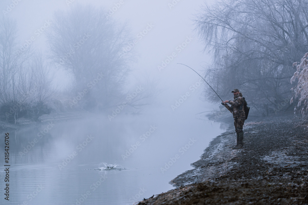 Fisher man fishing with spinning rod on a river bank at misty foggy winter, spin fishing, prey fishing