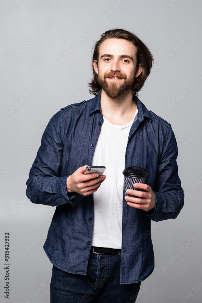 Portrait of a smiling young business man using smartphone and holding cup of coffee isolated on a gray background Portrait of a smiling young business man using smartphone and holding cup of coffee isolated on a gray background