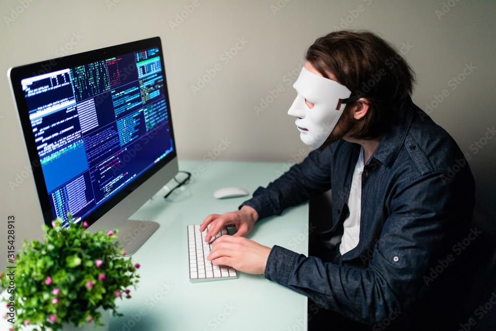 Anonymous computer hacker in white mask. Obscured dark face making silence gesture in Underground Secret Location Surrounded by Displays and Cables, darknet and cyber security concept.