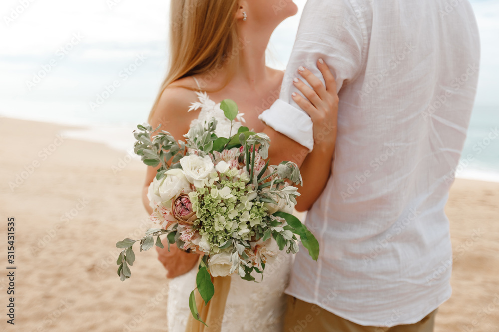 Φωτογραφία The bride hugs groom on the beach and holding bouquet of flowers