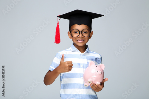 Young African American boy in graduation cap showing thumb up and holding piggybank on grey background
