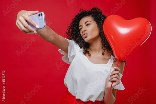 beautiful black woman making selfie on phone with heart shaped balloon isolated over red