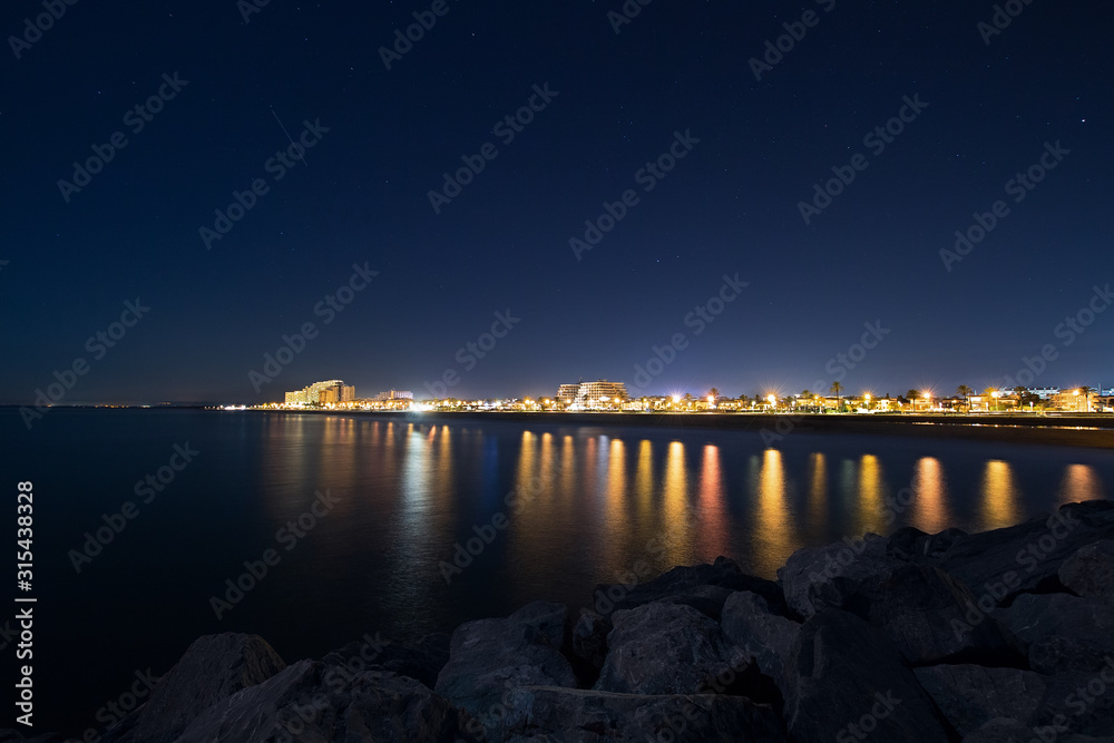 Night cityscape with sea and rocks in foreground and night star sky, night urban landscape of a Spanish city. Travel destination in Europe, card view with city lights and waterfront at night