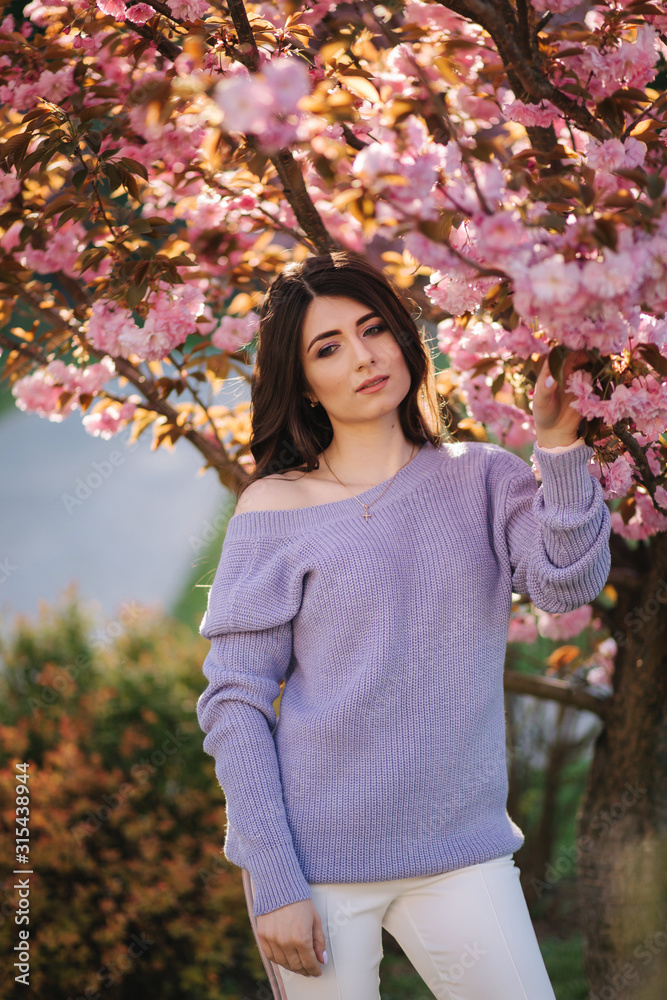 Attractive young woman in a purple sweater stand in front of sakura tree in the park Attractive young woman in a purple sweater stand in front of sakura tree in the park