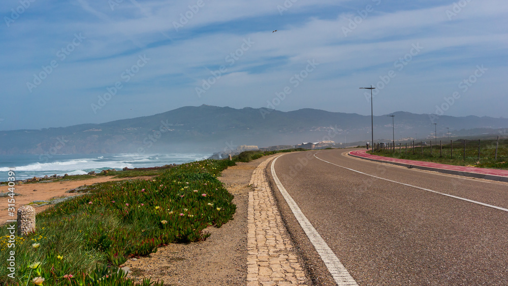 Empty asphalt road near the sea, leading towards the mountains on a hazy spring morning. Scenic coastal walk from Cascais to Guincho Beach in Portugal, one of the finest beaches in Lisbon region.