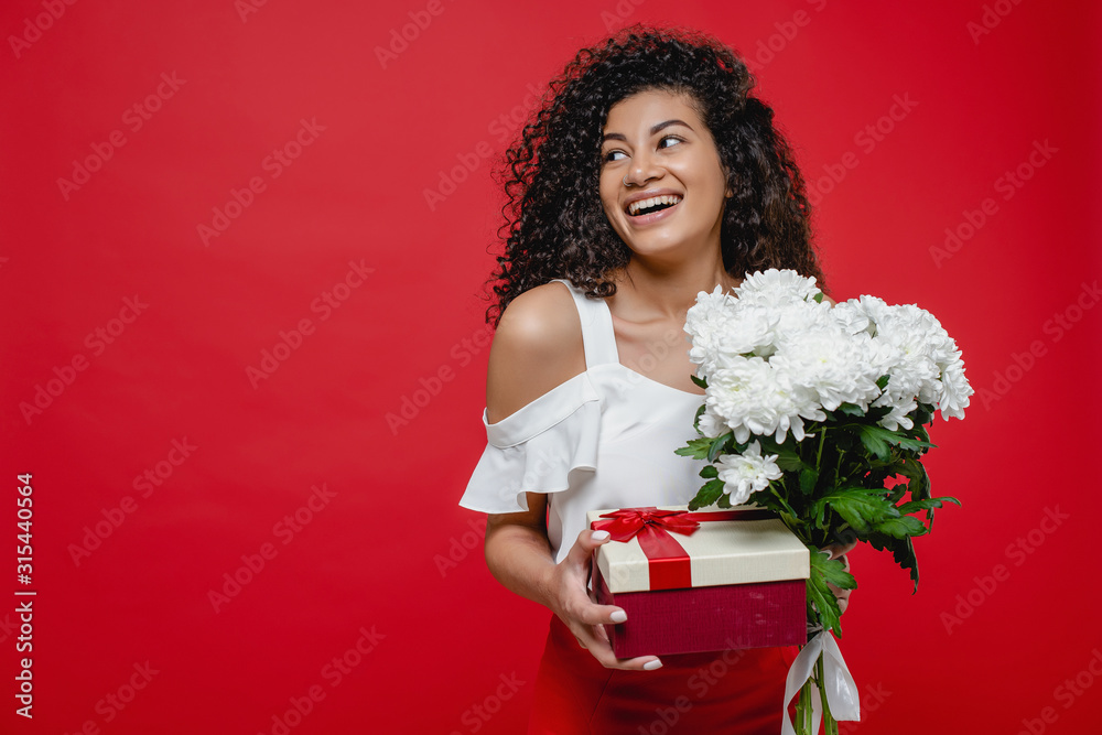 beautiful black woman smiling with gift box and bouquet of white flowers isolated over red background beautiful black woman smiling with gift box and bouquet of white flowers isolated over red background