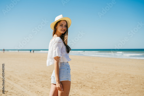 beautiful slim girl in a hat short shorts and a blouse stands on the sand by the ocean