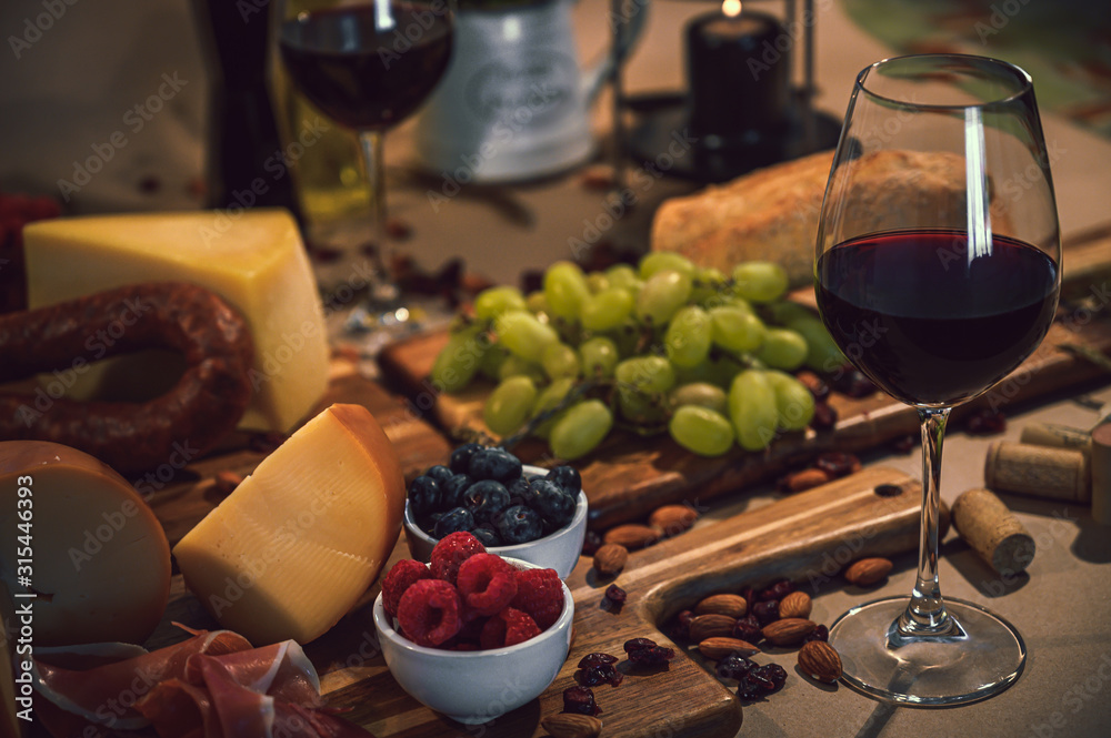 Glass of red wine accompanied by a variety of gourmet ingredients such as a variety of cheeses, cold meats, wine bottles, rustic bread and fruit on a rustic background