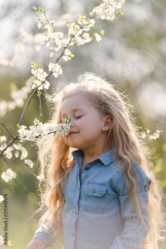 Little cute girl blonde with long hair sniffs a flowering tree branch in the park in spring.