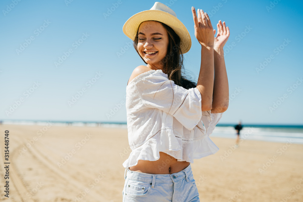 bright girl in a hat dancing clapping her hands, closing her eyes. behind the ocean and sand