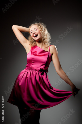 Young slim beautiful blond woman with curly hair in purple dress standing, smiling and looking at camera