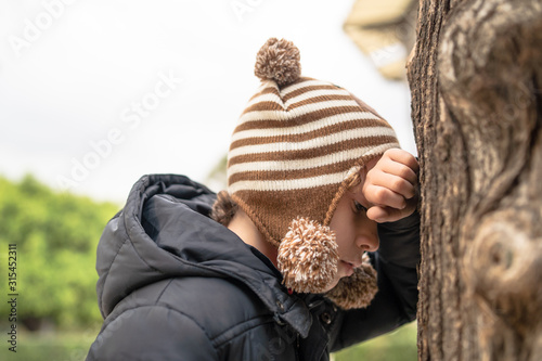 Cute kid playing hide and seek game. Hiding his face against a three in the city park.