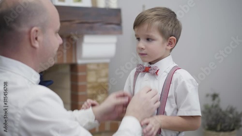 Close-up of confident Caucasian man helping little boy to tie a bow tie. Young father helping son to get ready for birthday party. Care, unity, support. photo