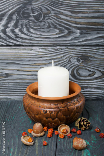 A large candle in a clay vessel. Acorns and mountain ash are scattered nearby. Standing on brushed boards.