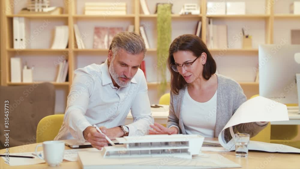 Architects with model of house sitting at the desk indoors in office, working. Architects with model of house sitting at the desk indoors in office, working.