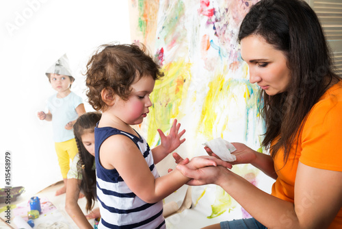 Woman with two babies painting with paints and brushes while sitting at childrens table, colorful background