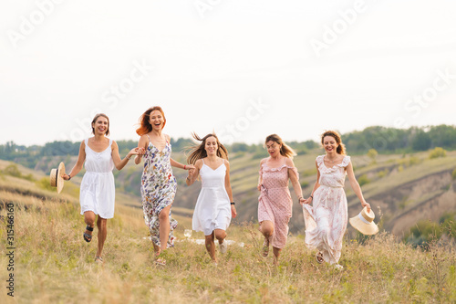 The company of cheerful female friends have a great time together on a picnic in a picturesque place overlooking the green hills. Girls in white dresses dancing in the field
