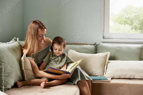 Young mother, read a book to her child, boy in the living room of their home, rays of sun going through the window