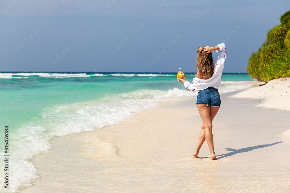 Blonde girl in denim shorts with fresh coconut in hand, on beach ocean in Maldives. Summer vacation travel. Banner panorama landscape. Blonde girl in denim shorts with fresh coconut in hand, on beach ocean in Maldives. Summer vacation travel. Banner panorama landscape.