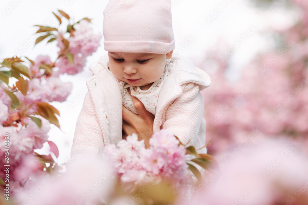 Cute baby girl between pink branches play with leaflets. Happy little girl surrounded by sakura Cute baby girl between pink branches play with leaflets. Happy little girl surrounded by sakura