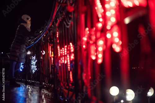 beautiful girl in a jacket, hat, shoes stands leaning on the railing of the stairs illuminated by new year's red lights in the night city