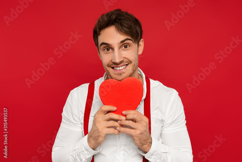 smiling handsome young man in suspender and shirt holding heart shaped box isolated on red, valentines concept © MarHo