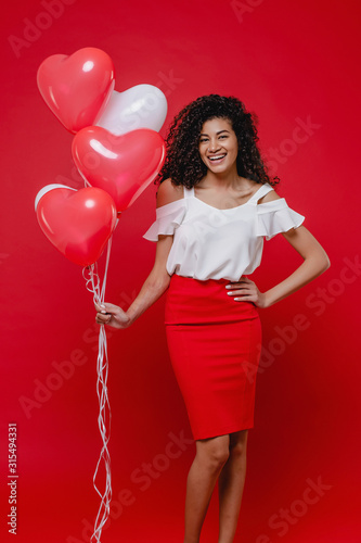 beautiful black woman smiling with heart shaped colorful balloons on red background © Anastasia