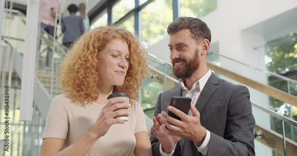 Hansome office worker in 30s showing funny photos to his female coworker at smartphone. Coworkers standing at hall with cofee cups, using mobilephone and looking at screen. People and tech. Hansome office worker in 30s showing funny photos to his female coworker at smartphone. Coworkers standing at hall with cofee cups, using mobilephone and looking at screen. People and tech.