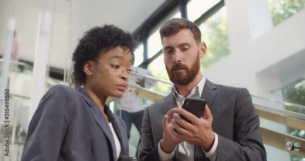 Male and female coworkers discussing current project and looking at smartphone screen. Corporate team member standing and talking while using smartphone in multi ethnic office. Male and female coworkers discussing current project and looking at smartphone screen. Corporate team member standing and talking while using smartphone in multi ethnic office.