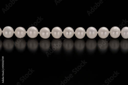 A strand of white round pearls lies horizontal across a black refective background. Low angle view of the pearls.