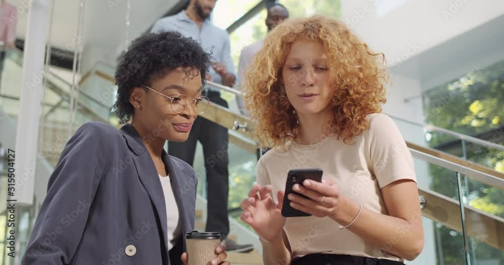 Girl manager showing photos to her female african friend coworker at smartphone. Women standing at hall with , using mobilephone and looking at screen during break. People and tech.