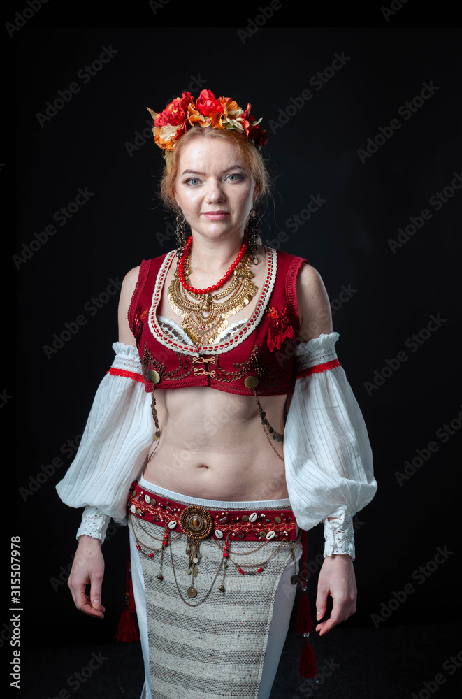 beautiful woman in traditional folk slavic costume for belly dance and trible with wreath, vest, skirt, sleeves and necklace standing and looking at camera on black background
