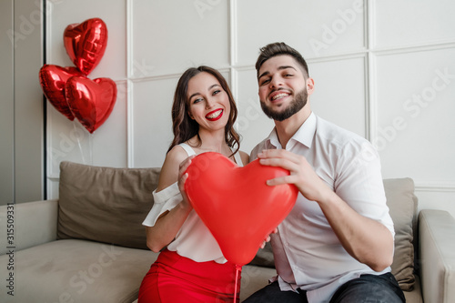 boyfriend and girlfriend with heart shaped balloon smiling and sitting on couch at home