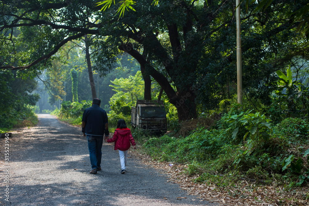 Back side of an Indian Bengali father and his little daughter with winter garments are walking through a pathway within a green natural forest in a winter morning. Indian lifestyle and winter.