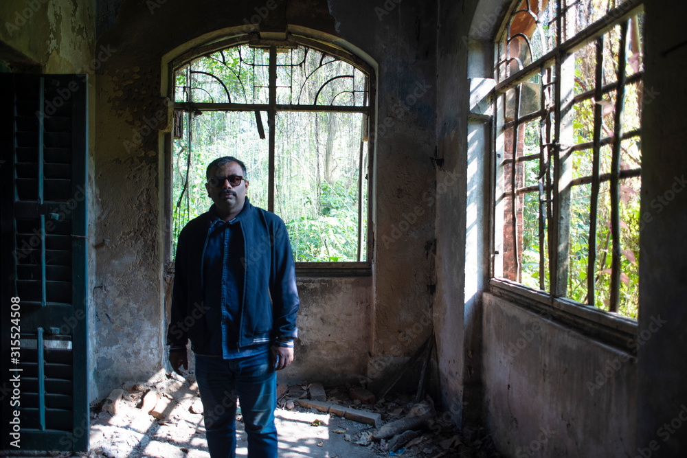 An Indian middle aged brunette man in winter clothing and sunglasses standing inside the ruins of an abandoned house with large windows. Vintage house and Indian lifestyle