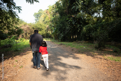 Back side of an Indian Bengali father and his little daughter with winter garments are walking through a pathway within a green natural forest in a winter morning. Indian lifestyle and winter.