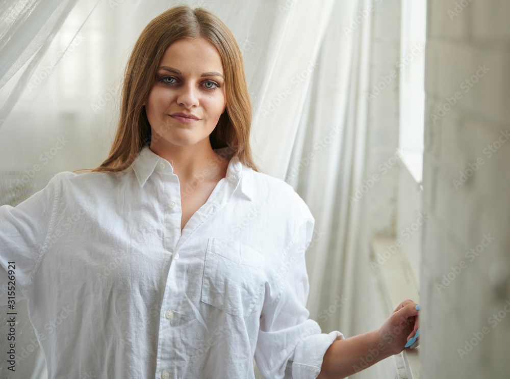 Stunning young Caucasian woman poses in studio wearing a men's white button-up shirt - near window