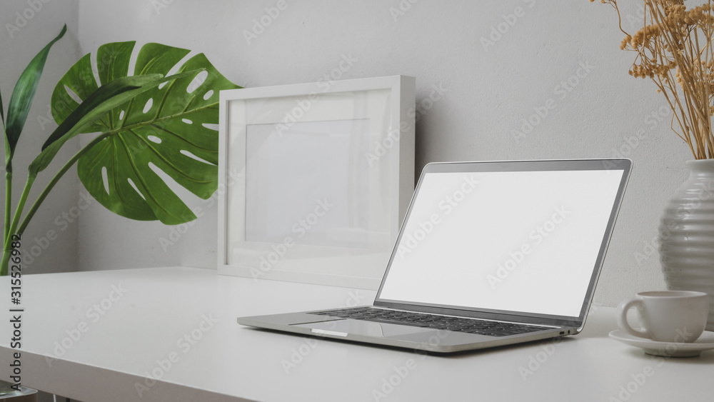 Fototapeta premium Close up view of workspace with blank screen laptop, mock up frame and ceramic vase on withe desk with white wall